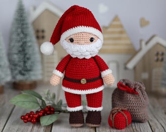 May include: A crocheted Santa Claus doll wearing a red suit with white trim, a white beard, and brown boots. The doll is holding a red gift bag and is standing in front of a wooden background with miniature houses and a Christmas tree.