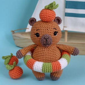 Könnte beinhalten: Ein gehäkeltes Capybara-Spielzeug in Braun mit einer kleinen Orange auf dem Kopf, dunkelbraunen Ohren und Füßen. Das Capybara trägt einen gestreiften Schwimmring in Orange, Weiß und Grün um die Taille und wird von zwei kleinen gehäkelten Orangen begleitet.