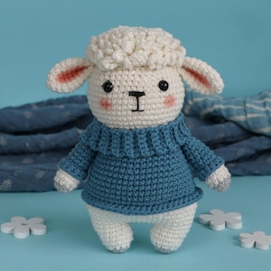May include: A crocheted sheep doll with a white, fluffy head and a blue sweater. The sheep has pink-lined ears, a white body, and grey hooves. The background is light blue with blue fabric and white flower decorations.