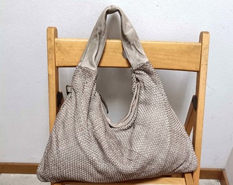 May include: A beige woven leather handbag with a slouchy design. The bag has two straps and is sitting on a wooden chair.