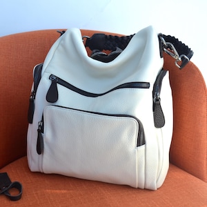 May include: White leather backpack with black accents and a braided strap. The bag has multiple zippered compartments and a flap closure.