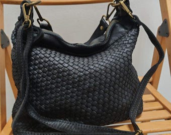 May include: A black woven shoulder bag featuring a textured, interlaced pattern across the front. The bag includes a long, adjustable strap with antique brass-toned metal hardware, including lobster claw clasps and a buckle for length adjustment.