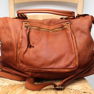 May include: A brown leather satchel bag with a zipped pocket on the front and a long adjustable strap. The bag is sitting on a wicker chair.