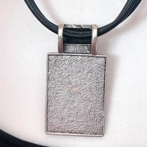 May include: A silver pendant with a textured surface hangs from a black leather cord. The pendant is rectangular in shape and has a smooth, polished edge.