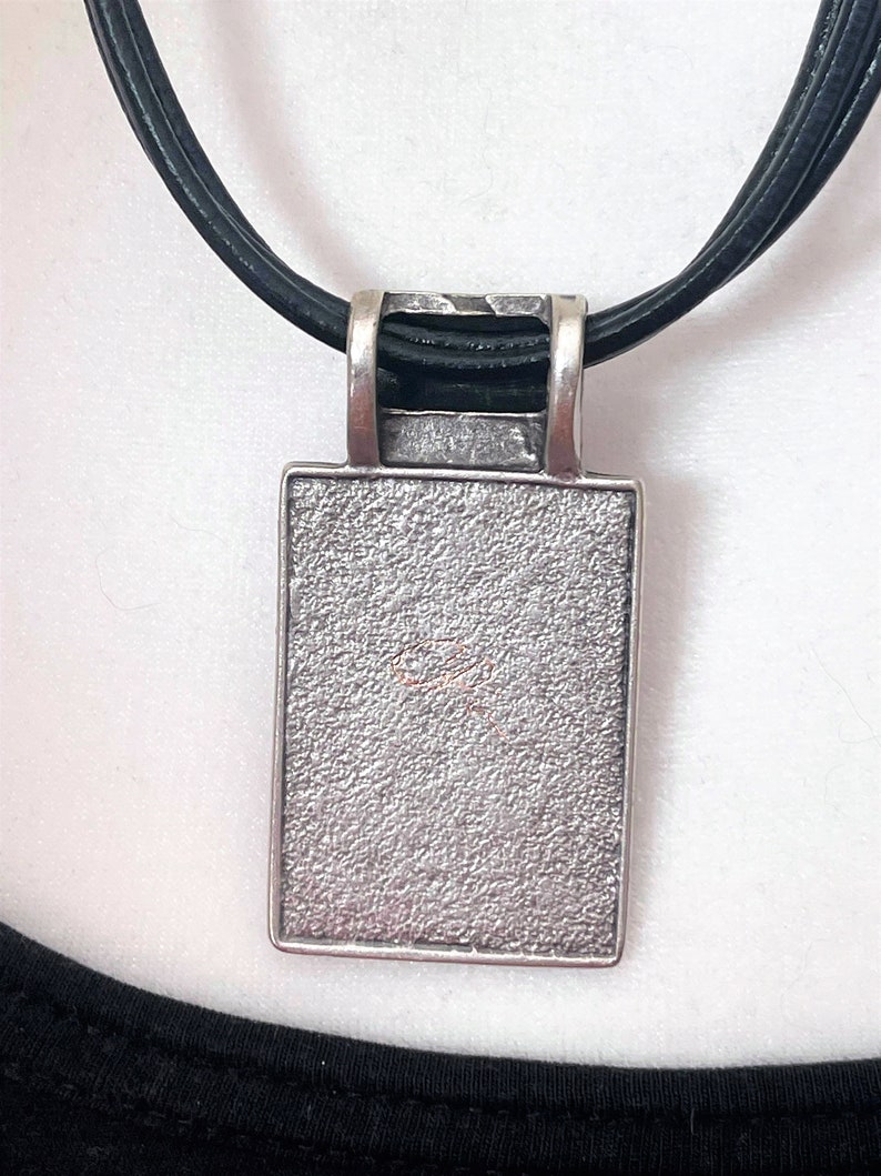 May include: A silver pendant with a textured surface hangs from a black leather cord. The pendant is rectangular in shape and has a smooth, polished edge.