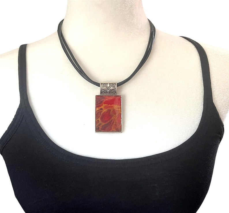 May include: A black leather cord necklace with a silver pendant featuring a red and orange abstract design.