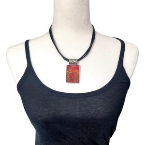 May include: A black leather cord necklace with a silver pendant featuring a red and orange abstract design.