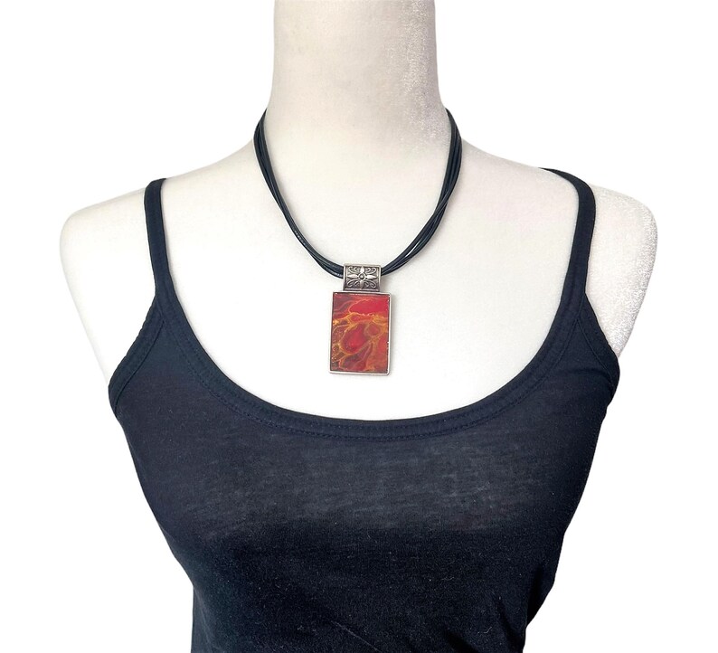 May include: A black leather cord necklace with a silver pendant featuring a red and orange abstract design.