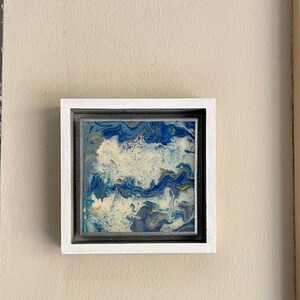 May include: A framed abstract painting with blue, white and yellow swirls. The painting is in a white frame with a black mount.