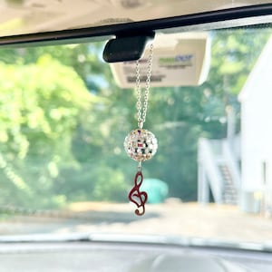 May include: A silver car charm with a disco ball and a treble clef hanging from the rearview mirror of a car.