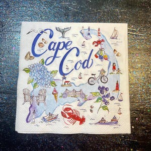May include: A square paper napkin featuring a map of Cape Cod with the text Cape Cod in blue script. The design includes illustrations of lighthouses, sailboats, a bicycle, a lobster, seashells, blue hydrangea flowers, and two wooden chairs.