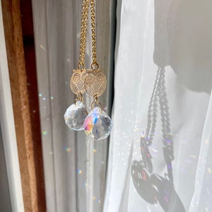 May include: A pair of gold owl charm necklaces with clear crystal drops hanging from them. The necklaces are hanging from a white curtain.