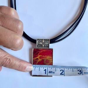 May include: A red and yellow abstract art pendant necklace with a silver frame and a black leather cord. The pendant measures 2.5 cm in length.