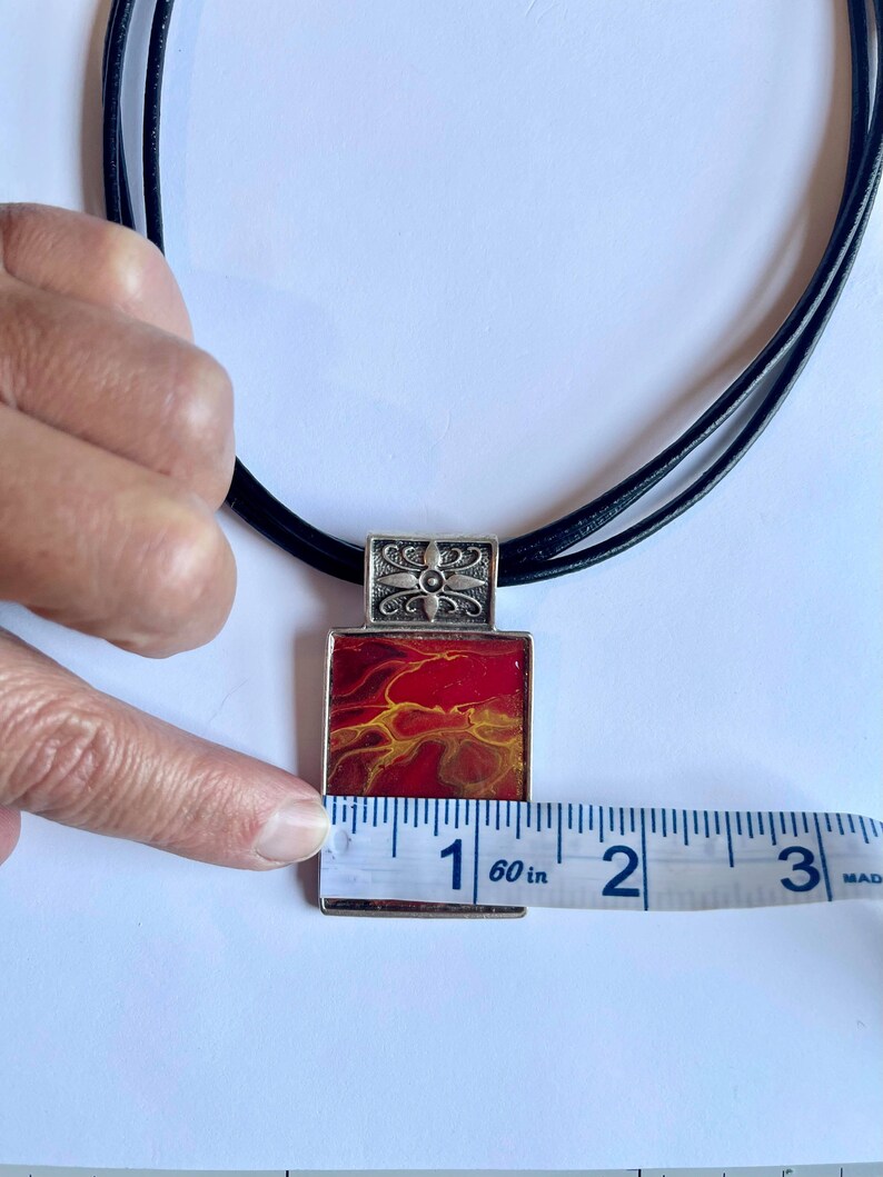 May include: A red and yellow abstract art pendant necklace with a silver frame and a black leather cord. The pendant measures 2.5 cm in length.