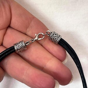 May include: A black leather cord necklace with silver-toned clasps and decorative end caps. The cord is flat and has a smooth texture.