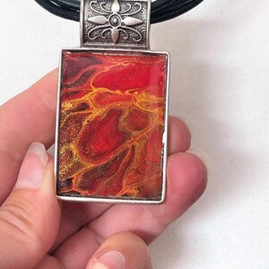 May include: A red and gold abstract pendant necklace with a silver floral design on the bail. The pendant is rectangular and has a black leather cord.
