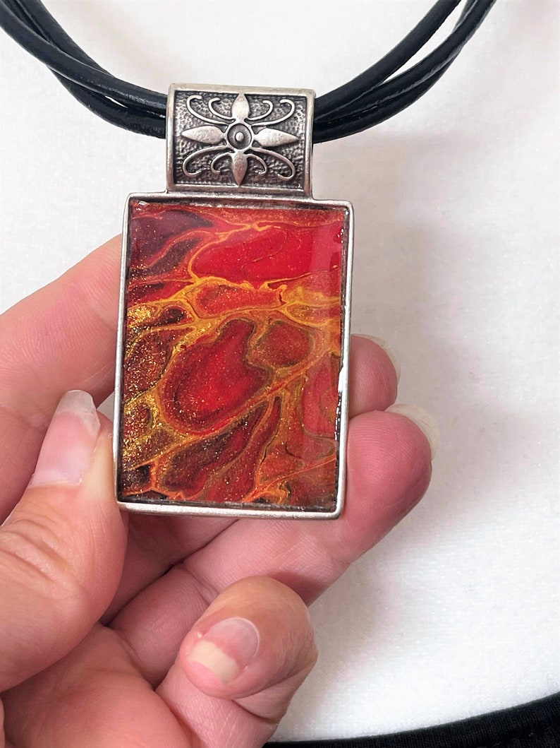 May include: A red and gold abstract pendant necklace with a silver floral design on the bail. The pendant is rectangular and has a black leather cord.