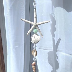 以下が含まれることがあります： 白いヒトデに薄い緑色のビーズがぶら下がっています。ヒトデはビーズと貝殻の紐に繋がっていて、茶色の貝殻と白い貝殻が含まれています。紐は白いカーテンからぶら下がっています。