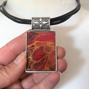 May include: A silver pendant necklace with a rectangular red, orange, and gold abstract resin pendant. The pendant is set in a silver frame and hangs from a black leather cord.