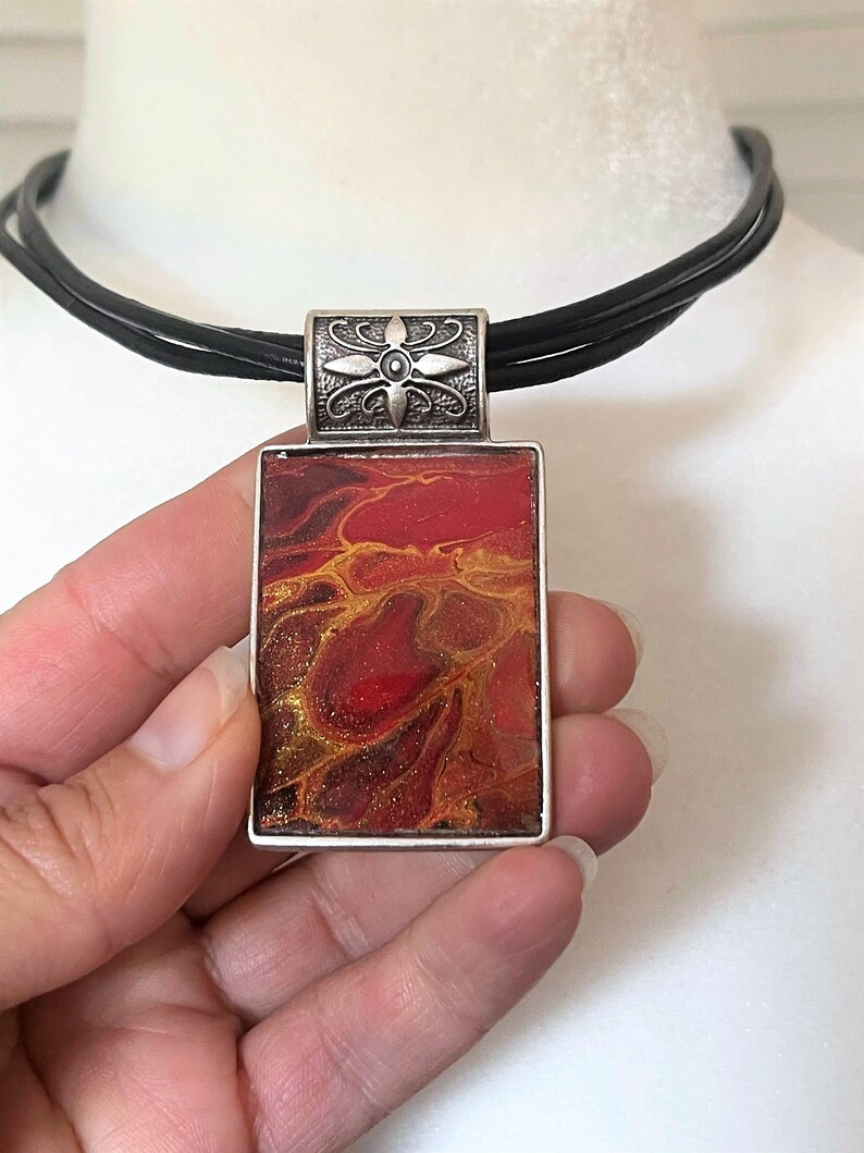 May include: A silver pendant necklace with a rectangular red, orange, and gold abstract resin pendant. The pendant is set in a silver frame and hangs from a black leather cord.