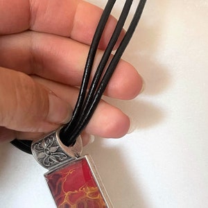 May include: A red and gold abstract pendant necklace with a silver filigree bail and a black leather cord.