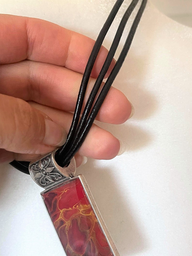 May include: A red and gold abstract pendant necklace with a silver filigree bail and a black leather cord.