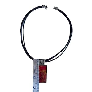 May include: A black leather cord necklace with a silver pendant featuring a red and orange abstract design. The pendant is approximately 5 cm long.
