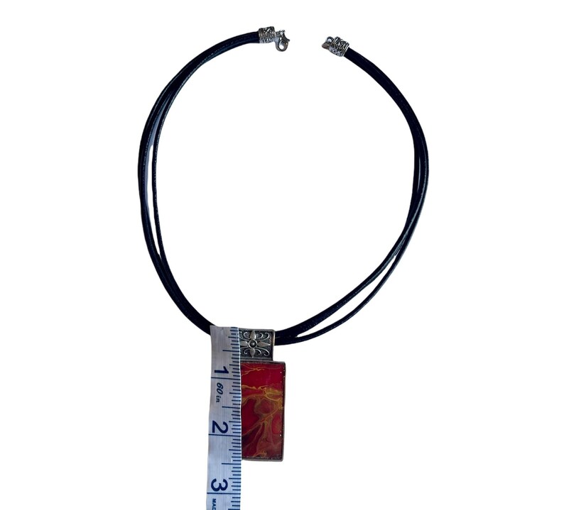 May include: A black leather cord necklace with a silver pendant featuring a red and orange abstract design. The pendant is approximately 5 cm long.
