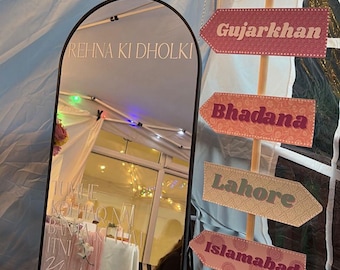 May include: A decorative mirror with the text "REHNA KI DHOLKI" and other text in a script font. Directional signs point to various locations, including Gujarkhan, Bhadana, Lahore, Islamabad, Rawalpindi, and Murree. The signs are pink and peach.