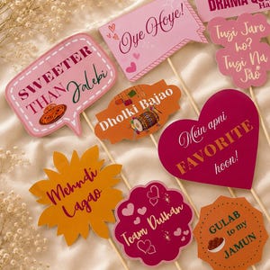 Puede incluir: Una colección de accesorios coloridos para fotomatón en palos de madera con varias formas y textos, incluyendo Sweeter Than Jalebi, Oye Hoye, Drama Queen, Tusi Jare ho, Dholki Bajao, Mein apni FAVORITE hoon, Mehndi Lagao, Team Dulhan y Gulab to my JAMUN.