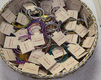 May include: A round woven basket filled with numerous thin, metallic bangles in various colors, including blue, green, pink, and silver. Each bangle is attached to a small, rectangular beige card printed with the text Maryam's Garden Party.