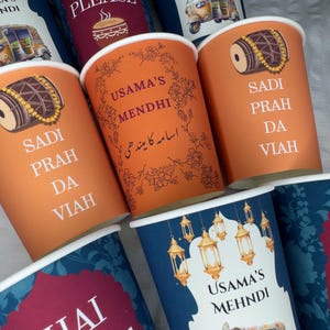 May include: A selection of paper cups in shades of orange, blue, and burgundy, each with unique designs and text. The cups display phrases such as "CHAI PLEASE", "USAMA'S MEHNDI", and "SADI PRAH DA VIAH". The cups are for a special event.