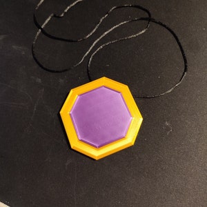 May include: A pendant necklace featuring a geometric, multi-layered design. The pendant has a purple center, surrounded by a yellow border. The necklace cord is black.