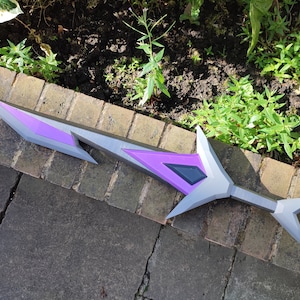May include: A purple and gray plastic sword with a sharp point and a jagged edge. The sword is lying on a brick surface.