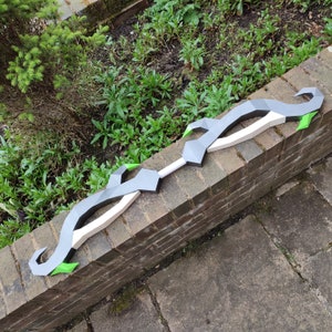 May include: A black and green bow and arrow prop with a white string. The bow is made of foam and has a sharp, pointed arrow.