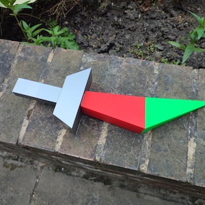 May include: A silver and red plastic toy sword with a green tip. The sword is lying on a brick surface.