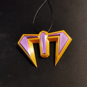 May include: A purple and gold 3D printed pendant shaped like a stylized letter M. The pendant is hanging from a black cord.