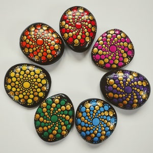 May include: Seven painted rocks with mandala designs. The rocks are painted in various colors, including red, yellow, blue, green, purple, and pink. Each rock has a gold dot pattern.