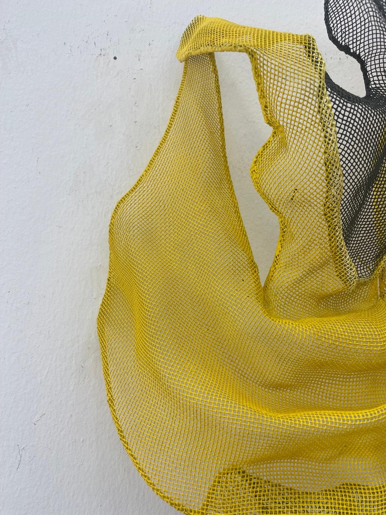 May include: A yellow mesh sculpture with a flowing, organic shape. The mesh is made of a fine, woven material and has a slightly textured surface.