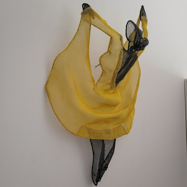 May include: A yellow and black wire sculpture of a dancer. The sculpture is made of wire mesh and is shaped like a dancer in a flowing dress. The sculpture is hanging on a white wall.