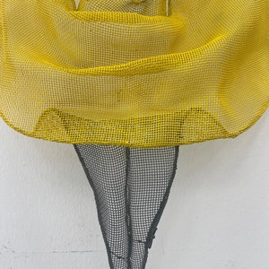 May include: A yellow mesh sculpture of a person with a flowing skirt. The sculpture is made of wire and is painted yellow. The sculpture is standing on a white background.