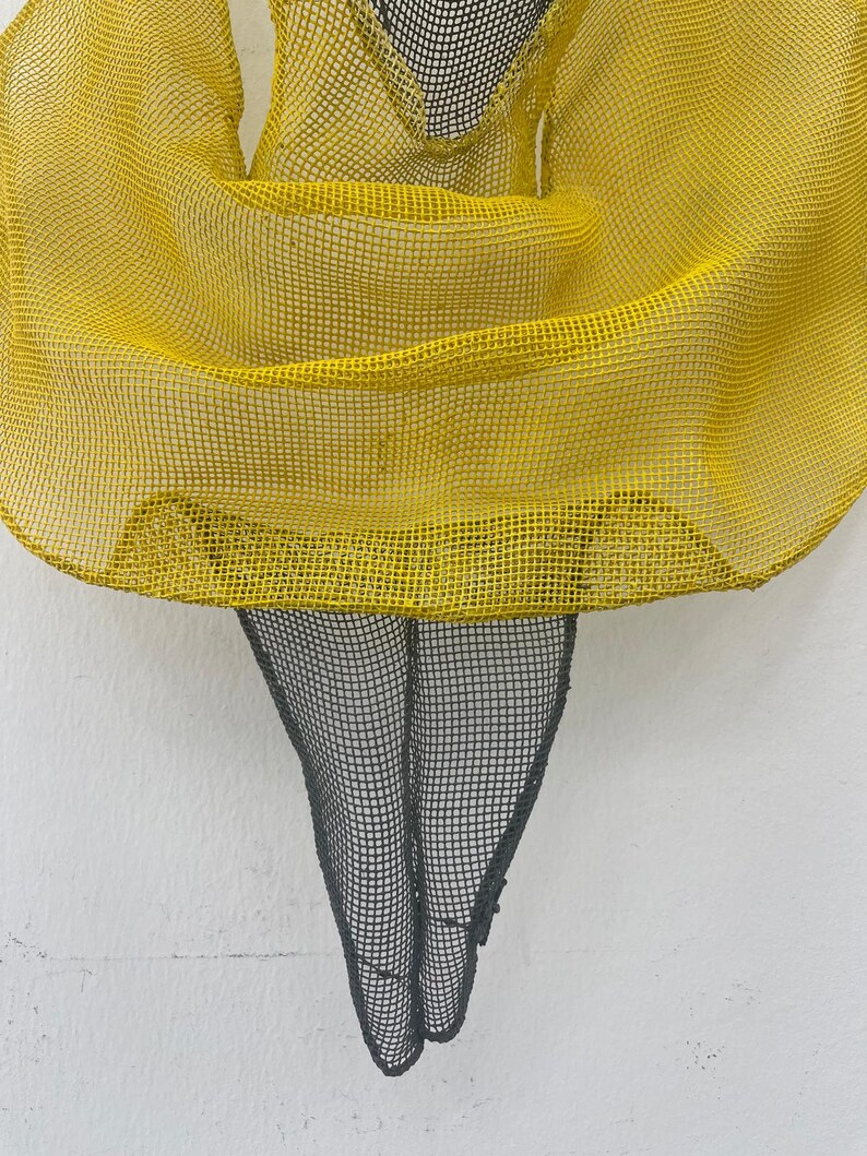 May include: A yellow mesh sculpture of a person with a flowing skirt. The sculpture is made of wire and is painted yellow. The sculpture is standing on a white background.