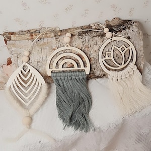 Könnte beinhalten: Drei Wandbehänge aus Holz mit Makramee-Fransen. Die Designs sind ein Regenbogen, eine Lotusblume und ein geometrisches Muster. Die Behänge sind mit Holzperlen aneinandergereiht.