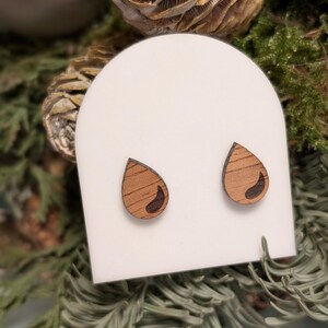 Op de afbeelding: Een paar houten oorbellen in de vorm van een traan met een donkerbruine houtnerf en een zwarte inleg. De oorbellen worden gepresenteerd op een witte kaart.