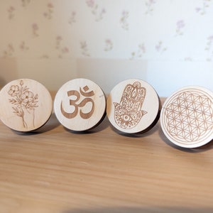 Könnte beinhalten: Vier Holz-Untersetzer mit verschiedenen Designs. Der erste Untersetzer zeigt eine Hand, die einen Blumenstrauß hält. Der zweite Untersetzer hat das Om-Symbol. Der dritte Untersetzer hat die Hamsa-Hand mit einem Auge in der Mitte. Der vierte Untersetzer hat ein Lebensblume-Design.