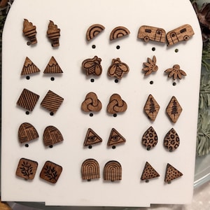 Op de afbeelding: Een wit displaybord met 20 paar houten oorbellen. De oorbellen zijn lasergesneden en hebben verschillende ontwerpen, waaronder ijshoorntjes, regenbogen, campers, bergen, bijen, bladeren, wolken, bomen, geometrische vormen, een oog en een luipaardprint.