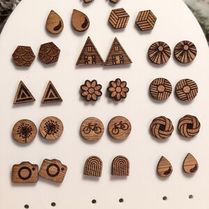 Op de afbeelding: Een verzameling houten oorstekers in diverse vormen en ontwerpen, waaronder bladeren, zeshoeken, driehoeken, cirkels, bloemen, fietsen, camera's, bogen en druppels. De oorbellen worden gepresenteerd op een witte standaard.