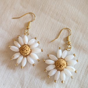 Könnte beinhalten: Ein Paar ohrringförmige Ohrringe. Jeder Ohrring hat einen zentralen, hellbraunen Stein, der von weißen, oval geformten Perlen umgeben ist, die Blütenblättern ähneln. Goldfarbene Perlen und Haken vervollständigen das Design, das auf einem cremefarbenen Stoff liegt.