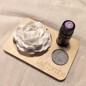 Op de afbeelding: Een houten diffuserbakje met de tekst "Duftprobe" erop gegraveerd. Het bakje heeft drie vakken: één voor een witte keramische bloem, één voor een klein grijs viltpad en één voor een klein flesje lavendelolie met het label "Laven-der".