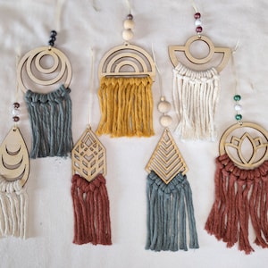 Könnte beinhalten: Sieben Wandbehänge aus Holz mit geometrischen Mustern und Makramee-Fransen. Die Muster umfassen einen Kreis, einen Halbmond, eine Raute, eine Blume, eine Sonne und zwei abstrakte Formen. Die Fransen sind in verschiedenen Farben, darunter Weiß, Braun, Rot, Gelb und Blau.